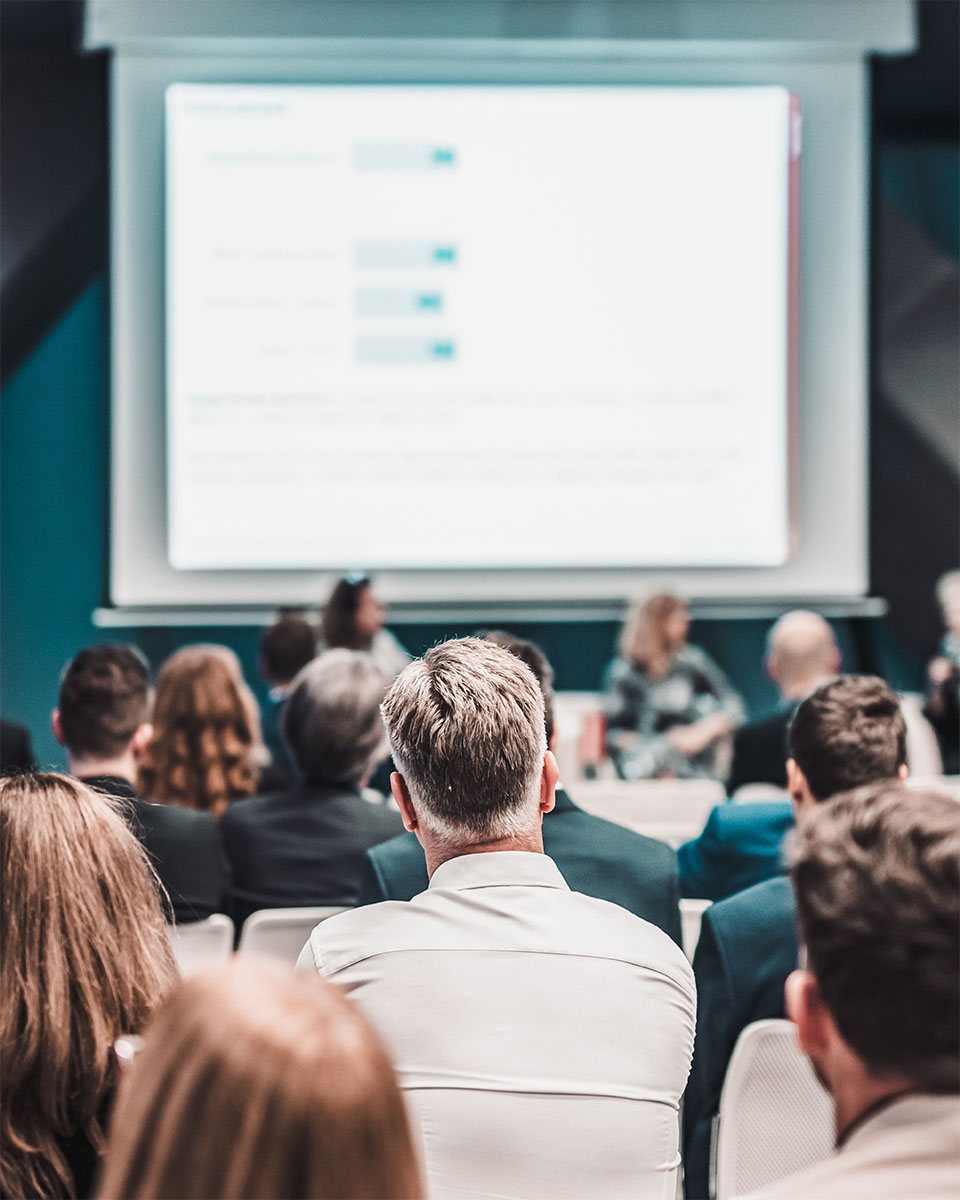 A group of professionals attends a private training session with a speaker presenting data on a large screen in a conference setting.