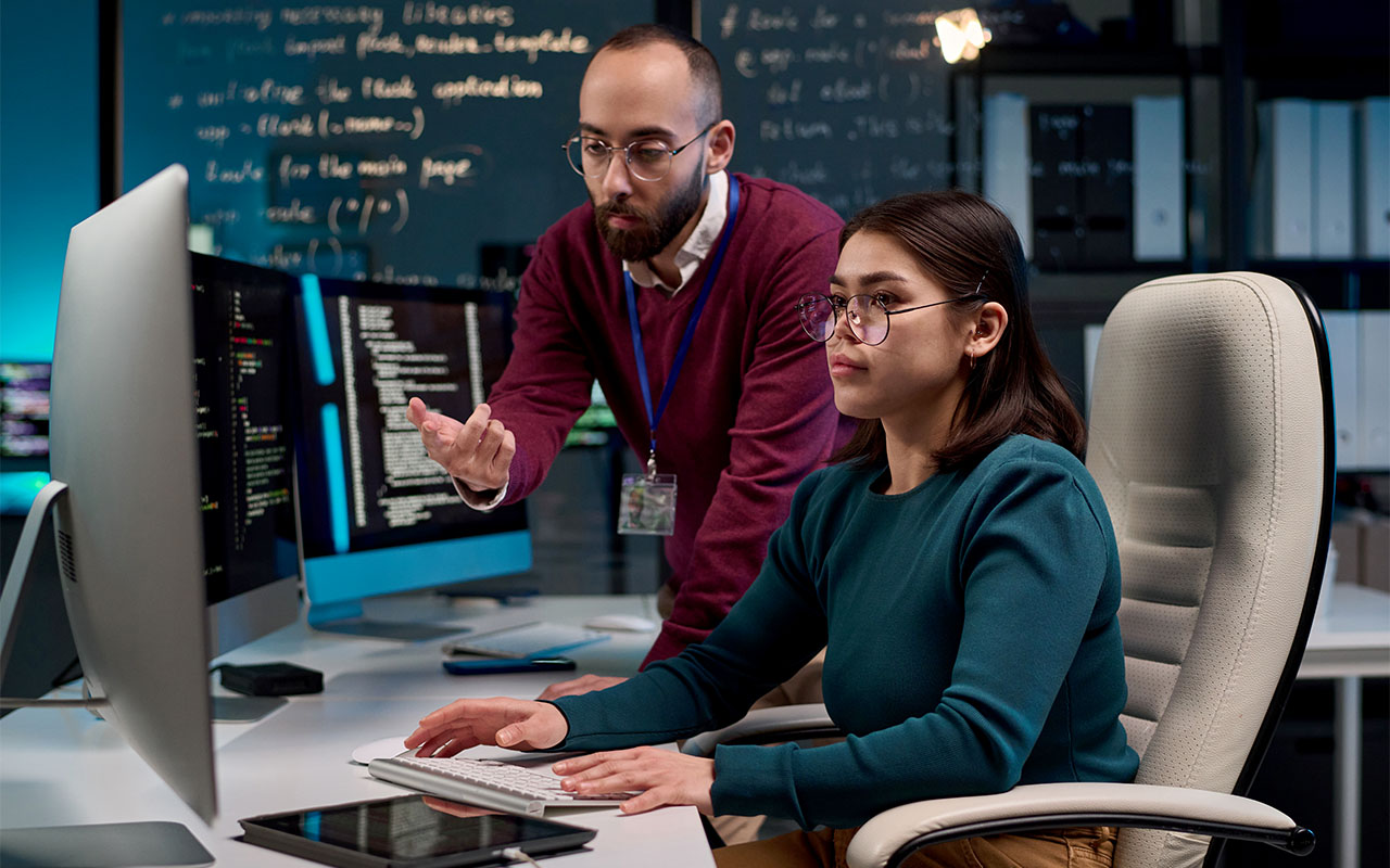 A mentor guides a programmer during private group training in a tech office, with code on screens and notes on a board in the background.